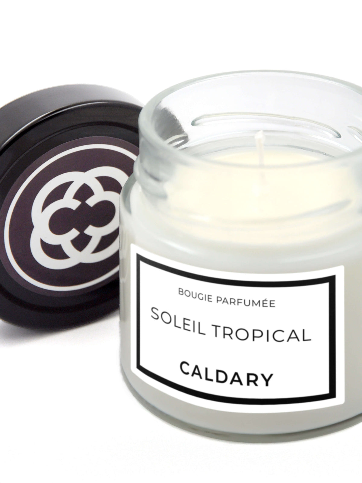 Sur fond blanc avec ombre portée. La bougie Soleil tropical révèle sa cire et sa mèche. À côté, debout sur sa tranche, le couvercle noir marqué du logo de l'atelier Caldary